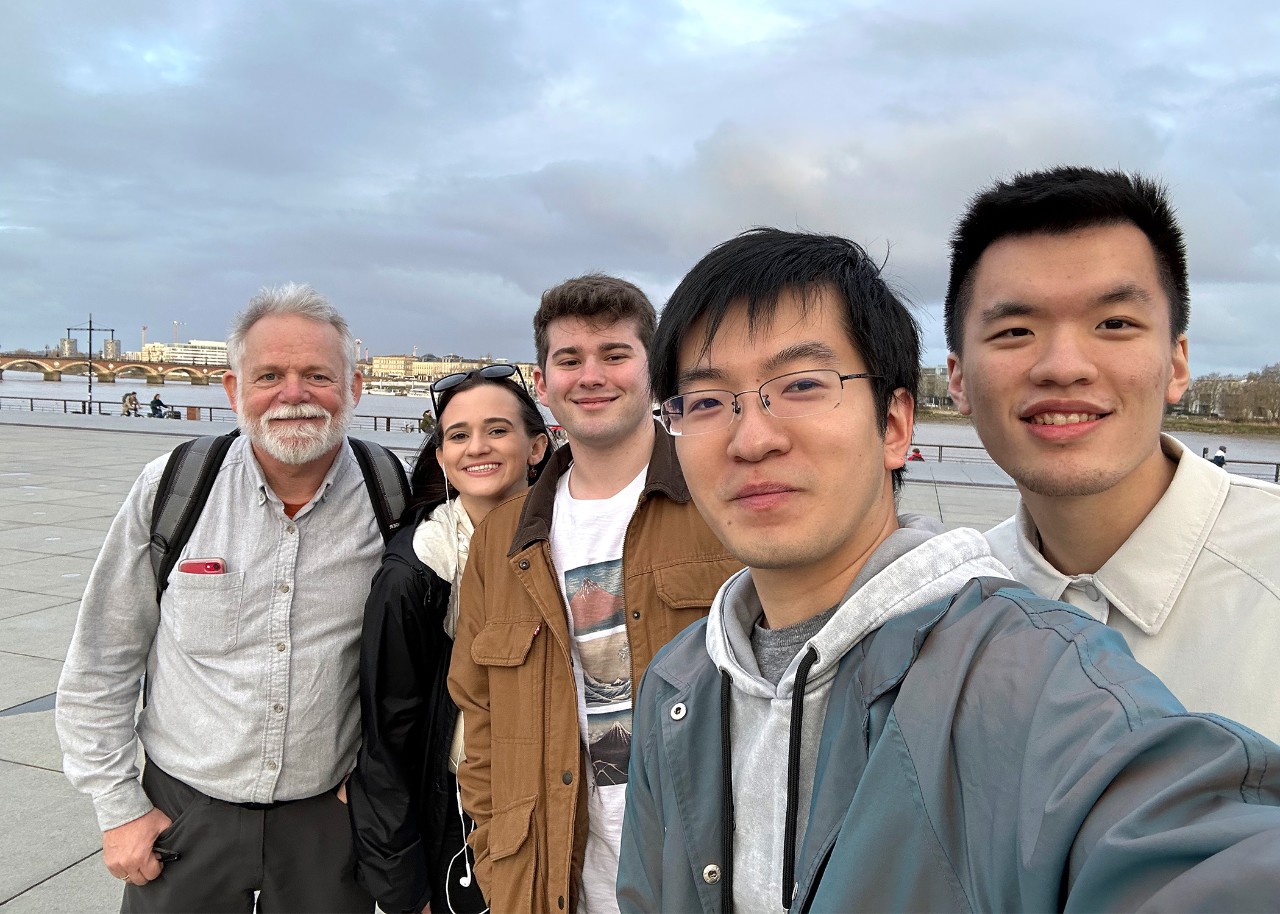 Selfie of a group of people; in the background are a river and bridge, with buildings further in the distance