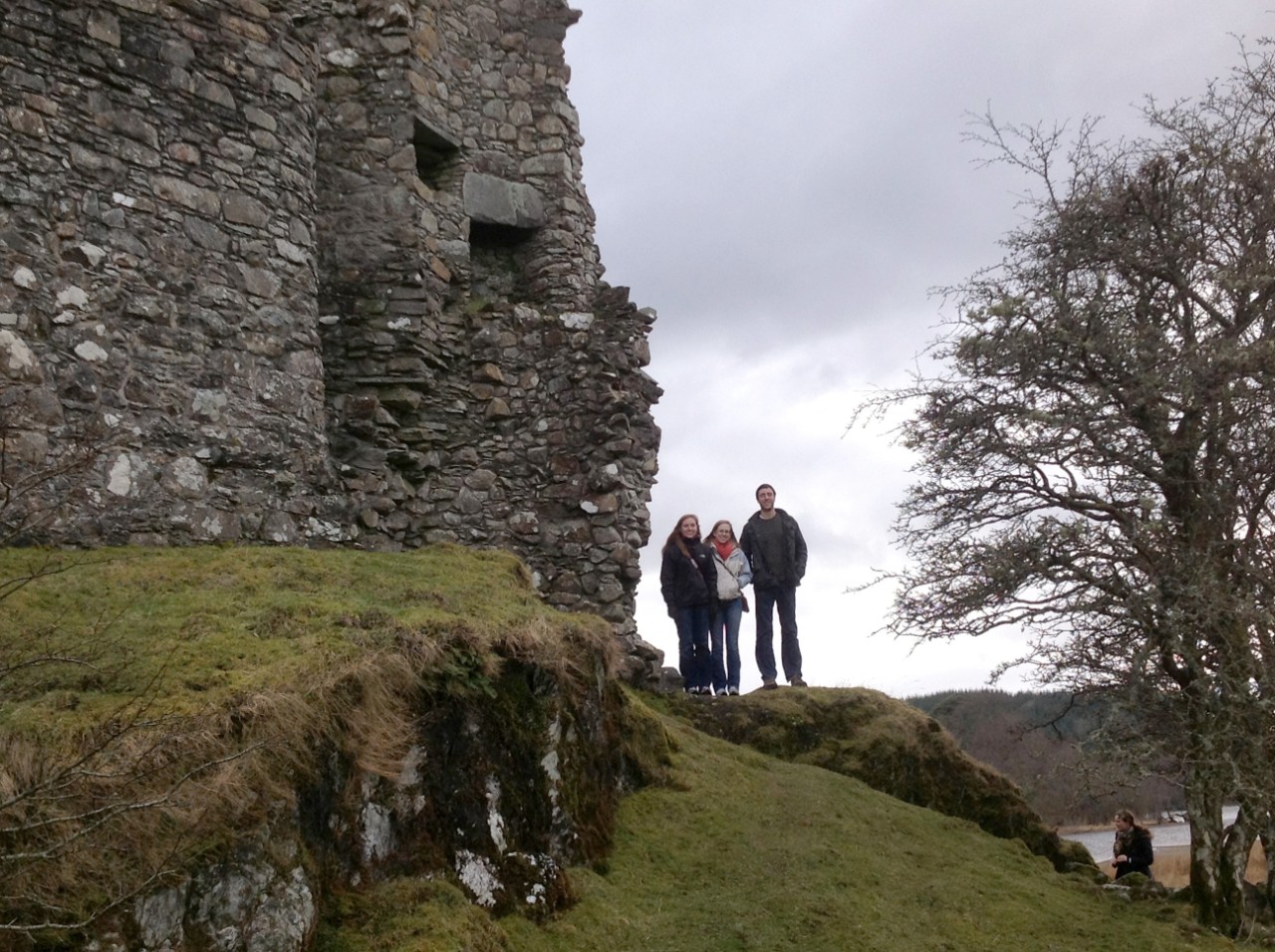 Three people stand on a chalky hill next to the worn wall of a stone fortress/castle in Scotland