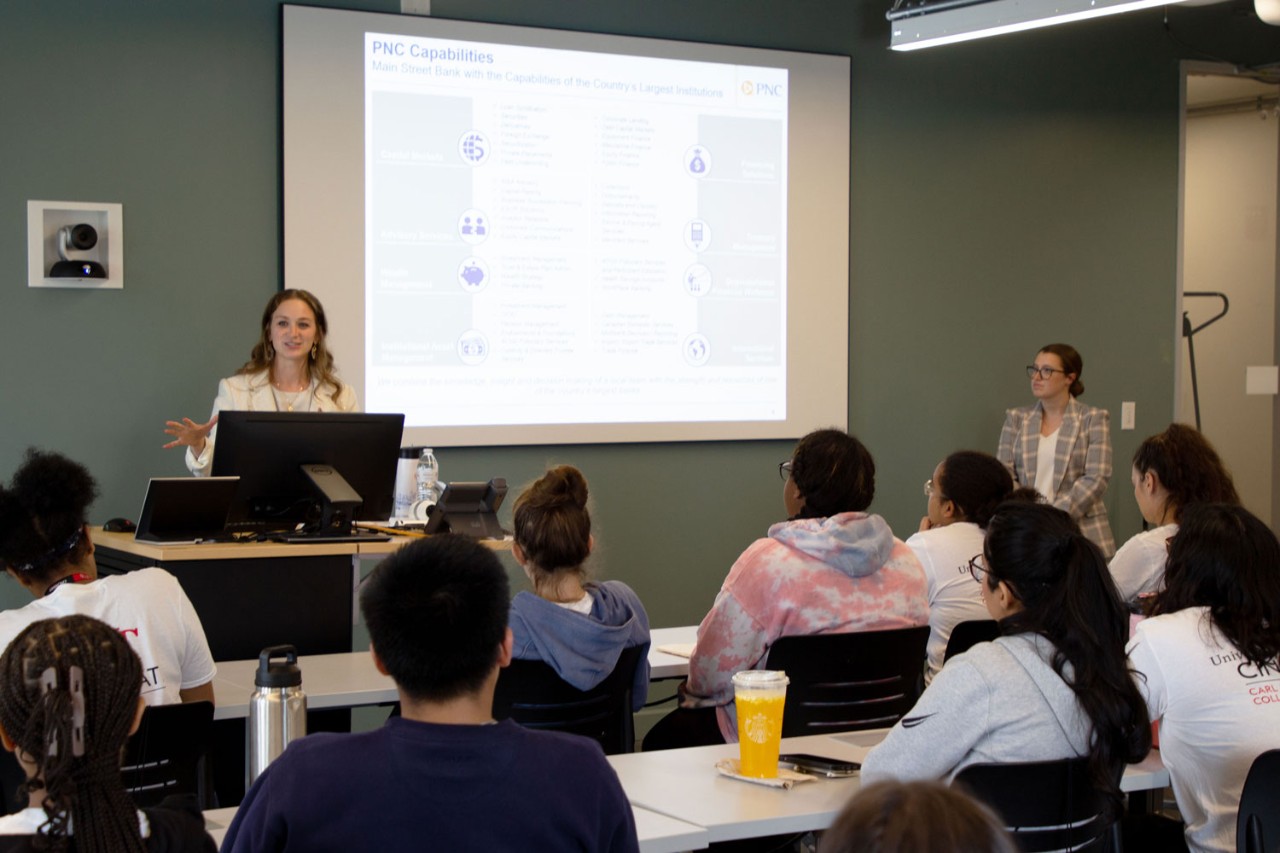 two women wearing professional attire stand in front of a classroom with a presentation on a white screen behind them