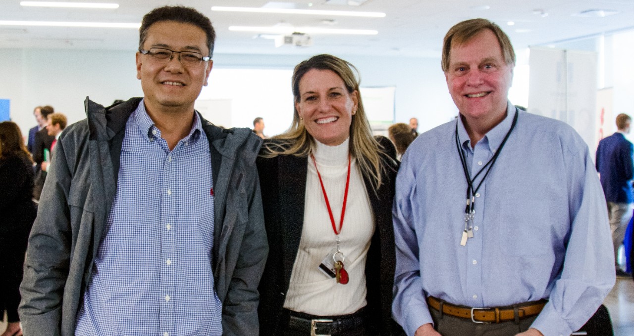 At an insurance career fair, two men in blue shirts, Seongho Song and Bob Higgins, take a picture with Ann Hoffman, in a white shirt and black jacket.