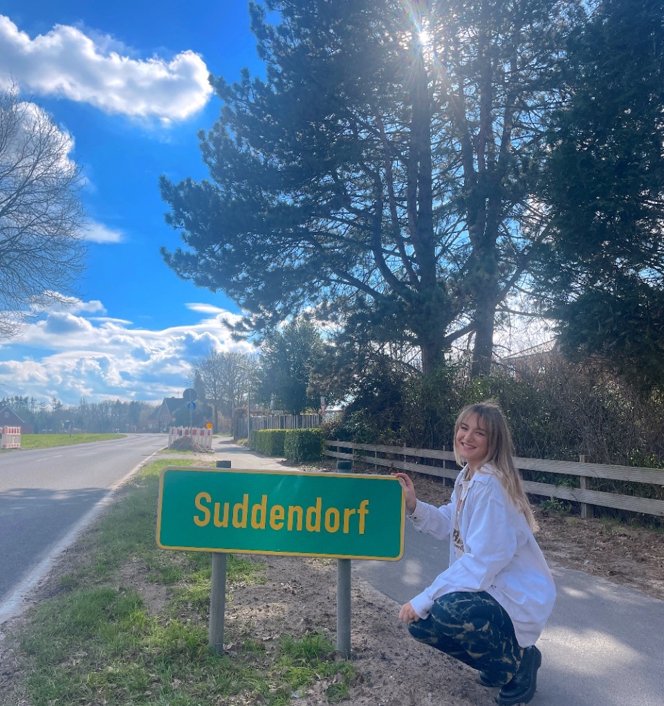 UC DAAP student Kathryn Suddendorf crouches down next to a road side that says "Suddendorf." 