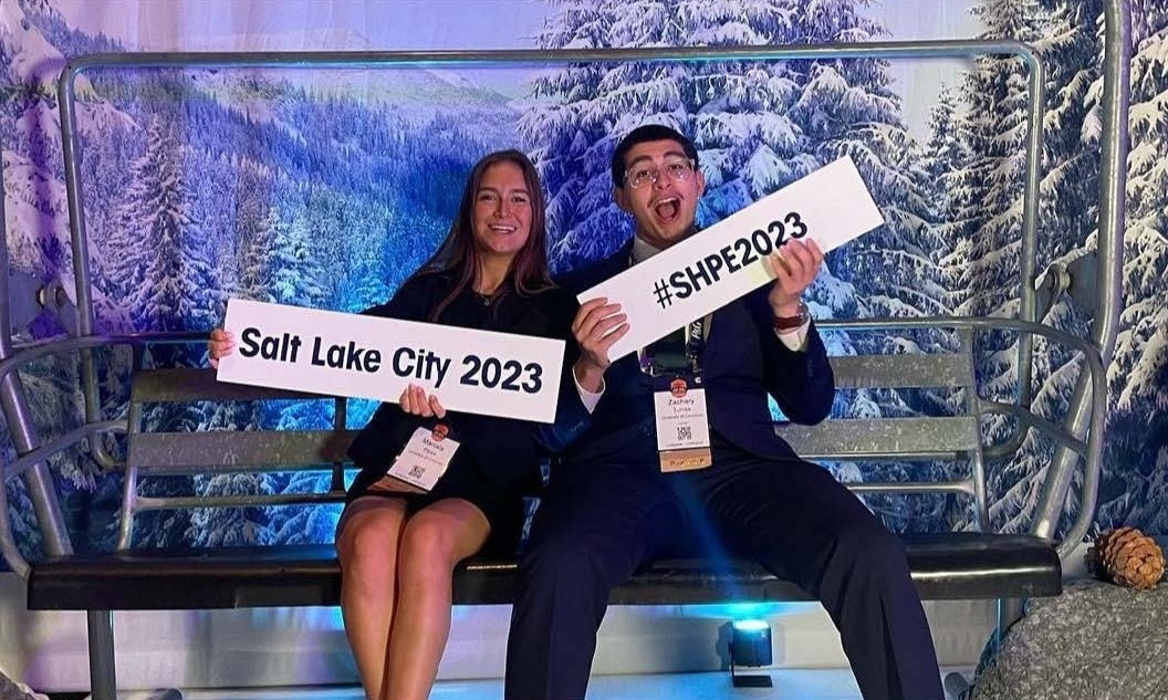 Marcela Palek (left) holds a sign that says "Salt Lake City 2023" and Zachary Torres (right) holds a sign that says "#SHPE2023"