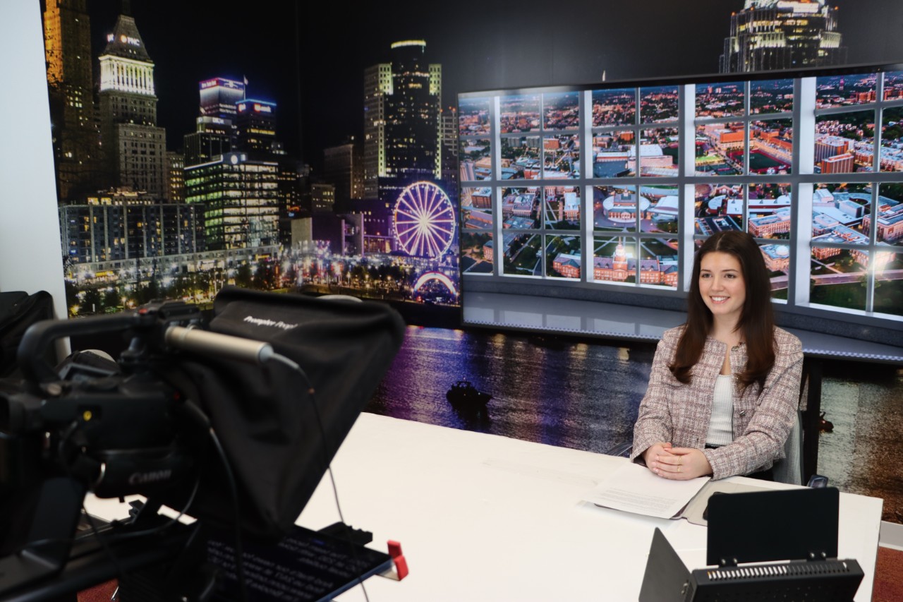 Student anchor poses in front of newscast background.