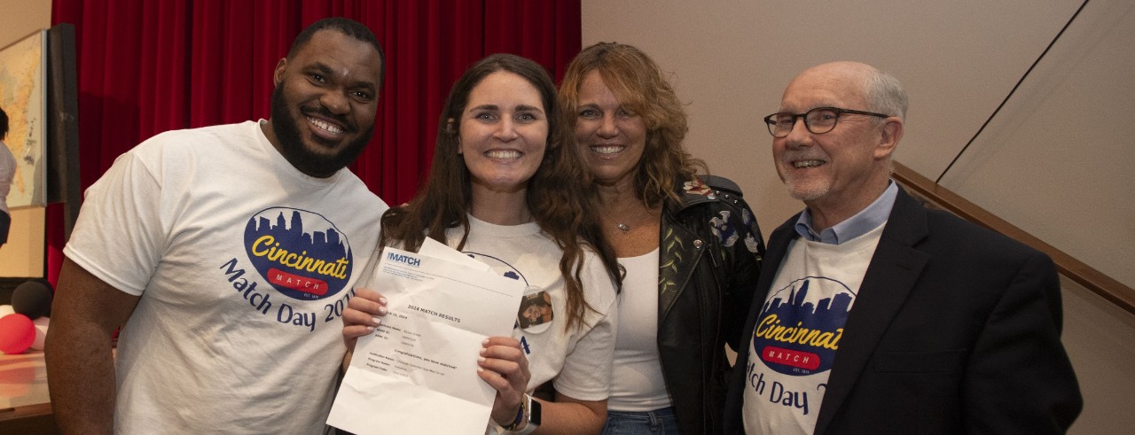 Two men and a woman stand and smile while another woman smiles and holds up her Match Day letter