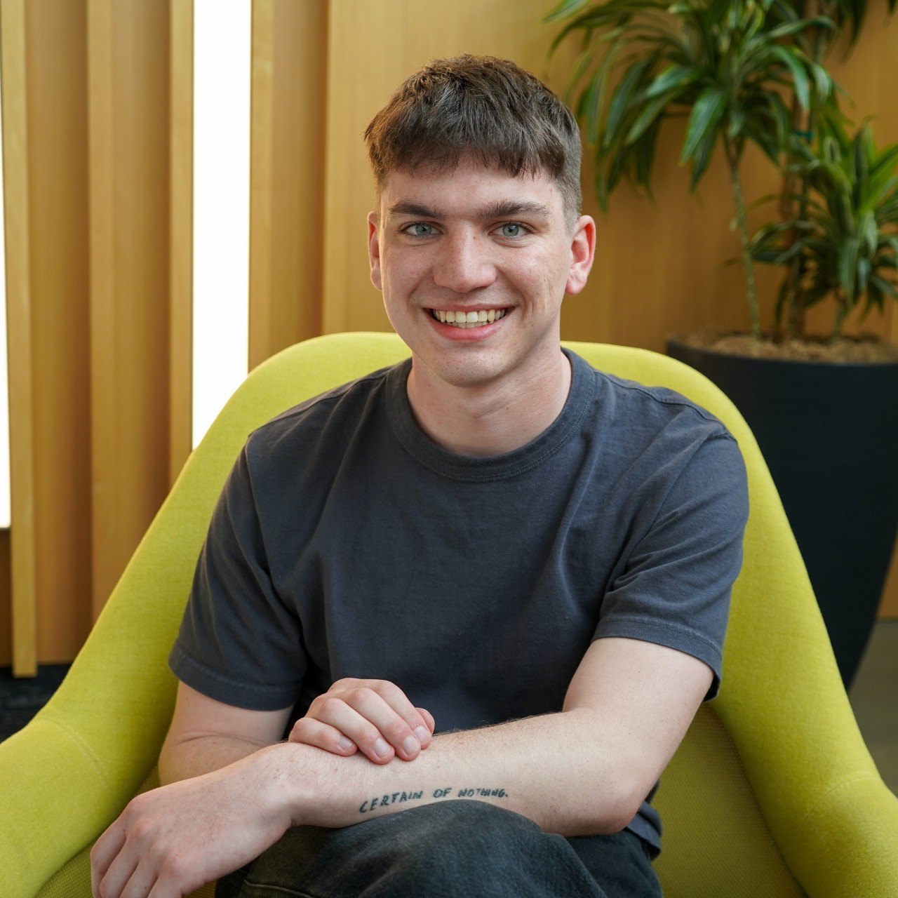 Headshot of UC student Max Kemats, he is smiling while sitting with his legs crossed on a bright green chair. His arms are crossed, showing a tattoo on his forearm that says "CERTAIN OF NOTHING."