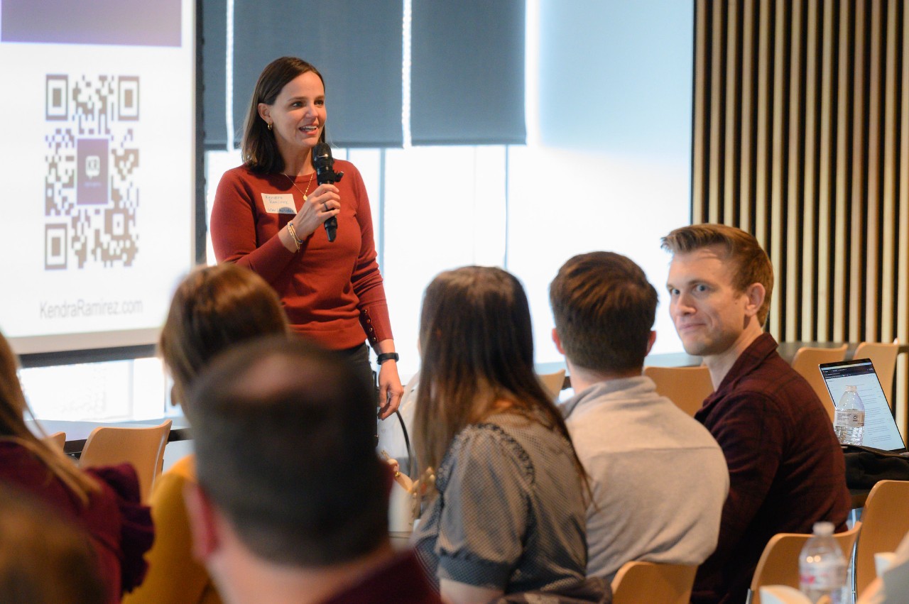 Kendra Ramirez, with a screen displaying a QR code behind her, holds a microphone and speaks to a room of people.