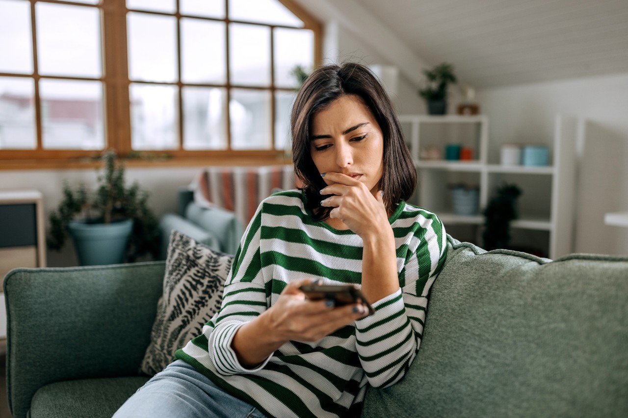 A woman looks concerned looking at her phone while scrolling on her couch.