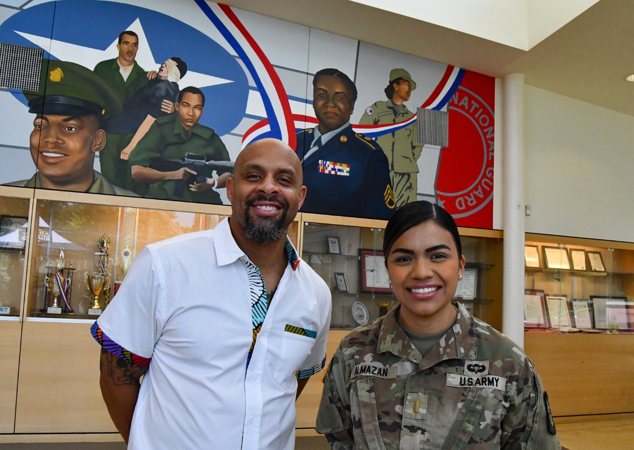 Two people stand in front of a mural featuring scenes from the U.S. military.