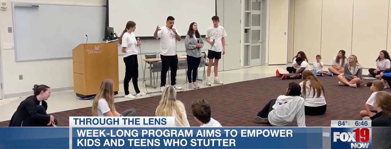 Camp staff members stand in front a room of kids and teens and model effective communication while speaking into microphones.