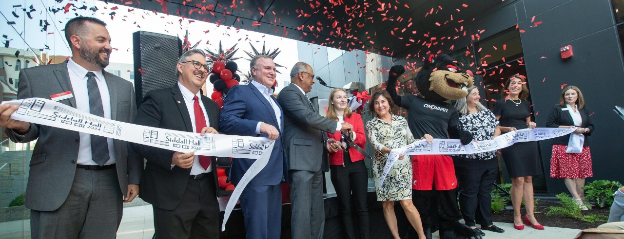 Ten UC administrators including President Pinto, the student body president Madison Wesley and the Bearcat stand and hold a ribbon for a ribbon cuting. Confetti cannon goes off.