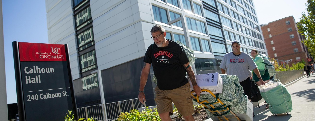 image of a father with UC T-shirt pulling a hand cart loaded with bags and boxes pass a Calhoun Hall sign