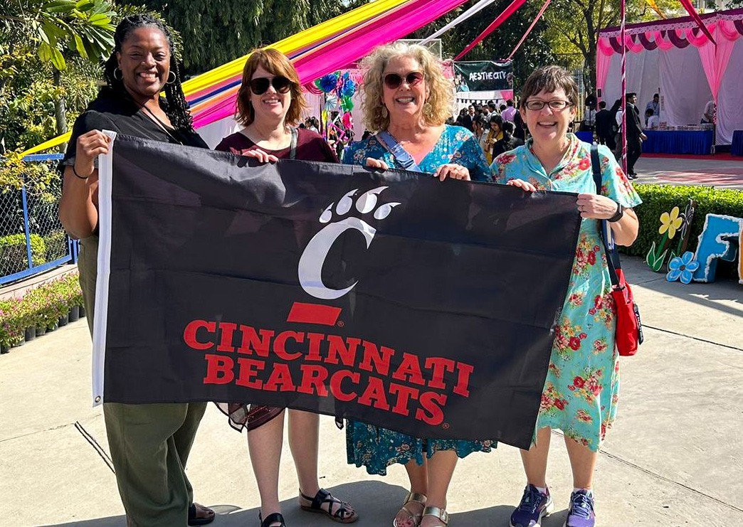 Four women hold up a banner that reads “Cincinnati Bearcats.” Behind them are colorful streamers and a decorated stage.