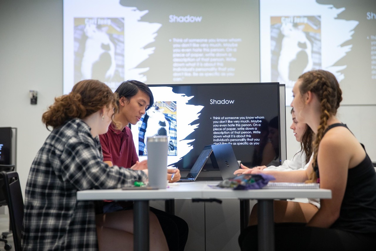 three students sit at a desk with large screen in front of them in a class