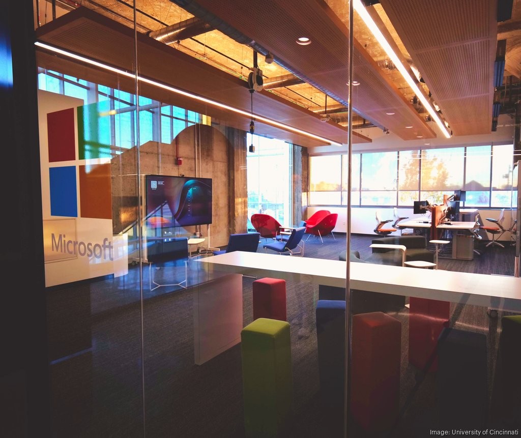 The Microsoft logo is on glass outside a space with tables, chairs, desks and a television screen.