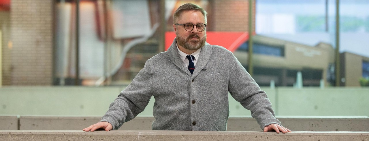 Portait of Jeffrey Blevins standing at a railing in an atrium with a glass window behind him.