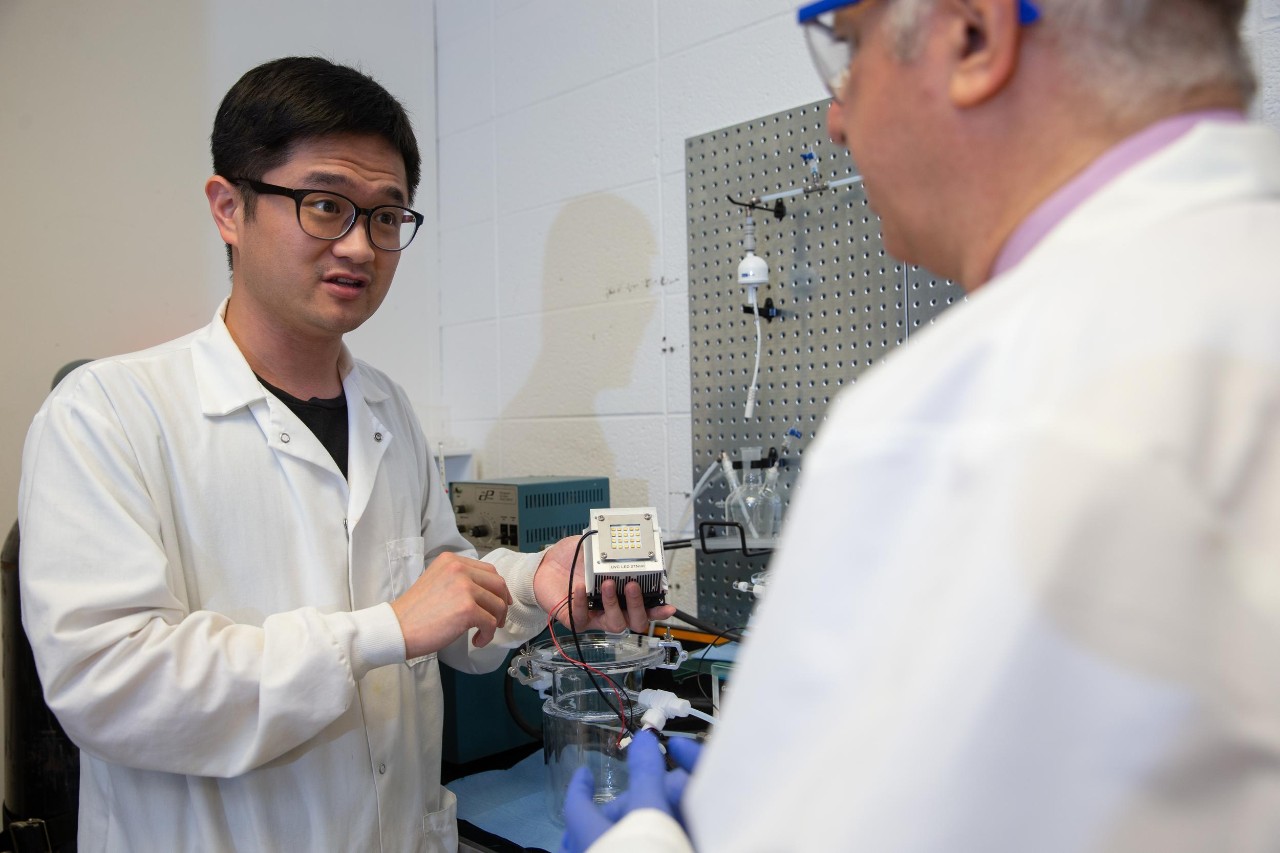 Minghao Kong in a labcoat holds up a sensor in front of Dionysios Dionysiou.