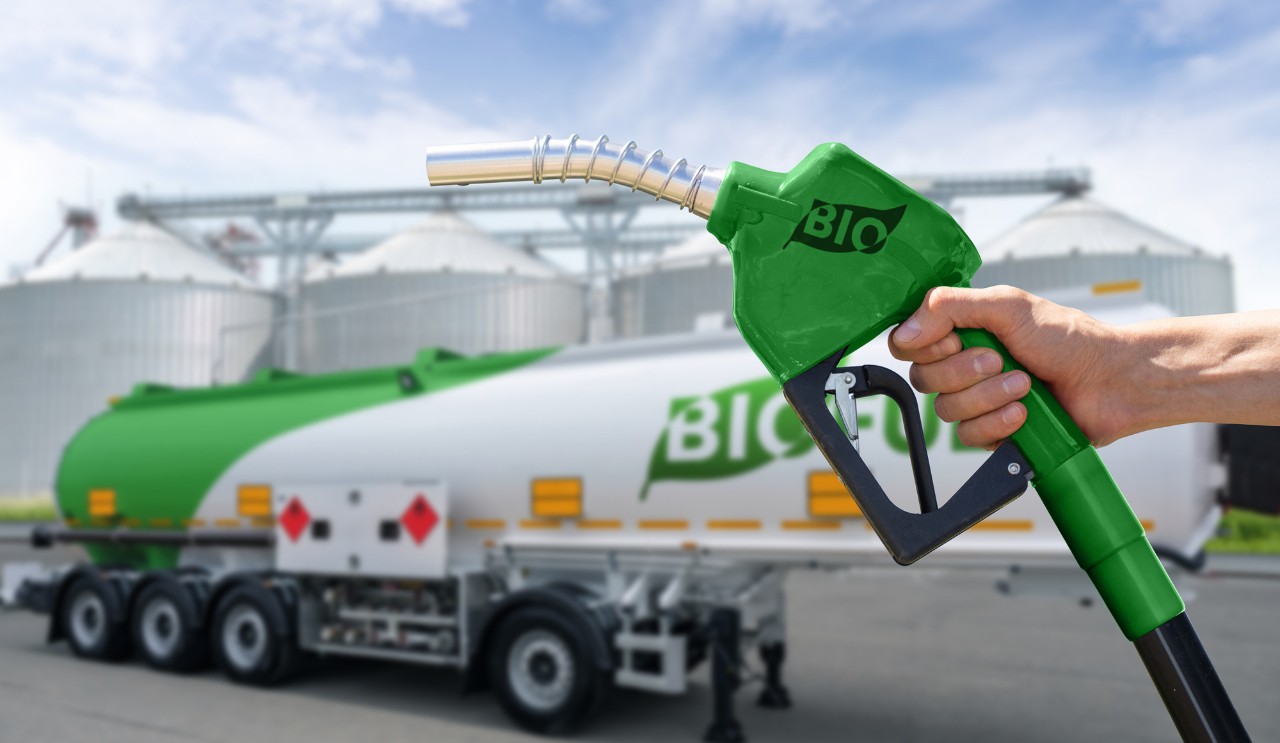 A hand holds a gas nozzle in front of a tanker truck that says biofuel.