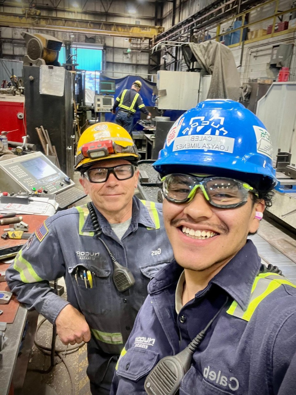 Caleb Loayza takes a selfie with another man while on co-op. They wear blue work uniforms and hard hats. 