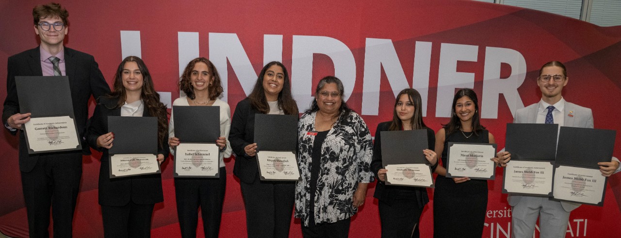 One professor and seven students holding awards certificates pose in front of a red banner with large letters reading 'Lindner'