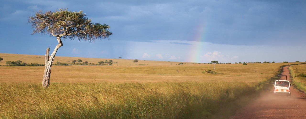 Masai Mara plains in Kenya with safari van on right and rainbow under blue skies