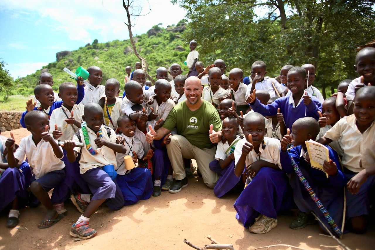 Several Tanzanian elementary students in white shirts and navy blue pants or skirts surround UC's Dr. Chris Lewis kneeling in center.
