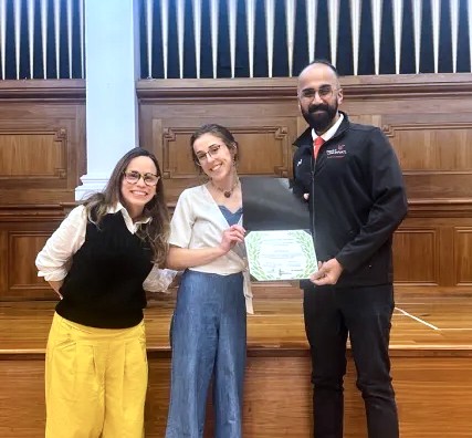 Two women on left stand with a man on right as he holds an award.