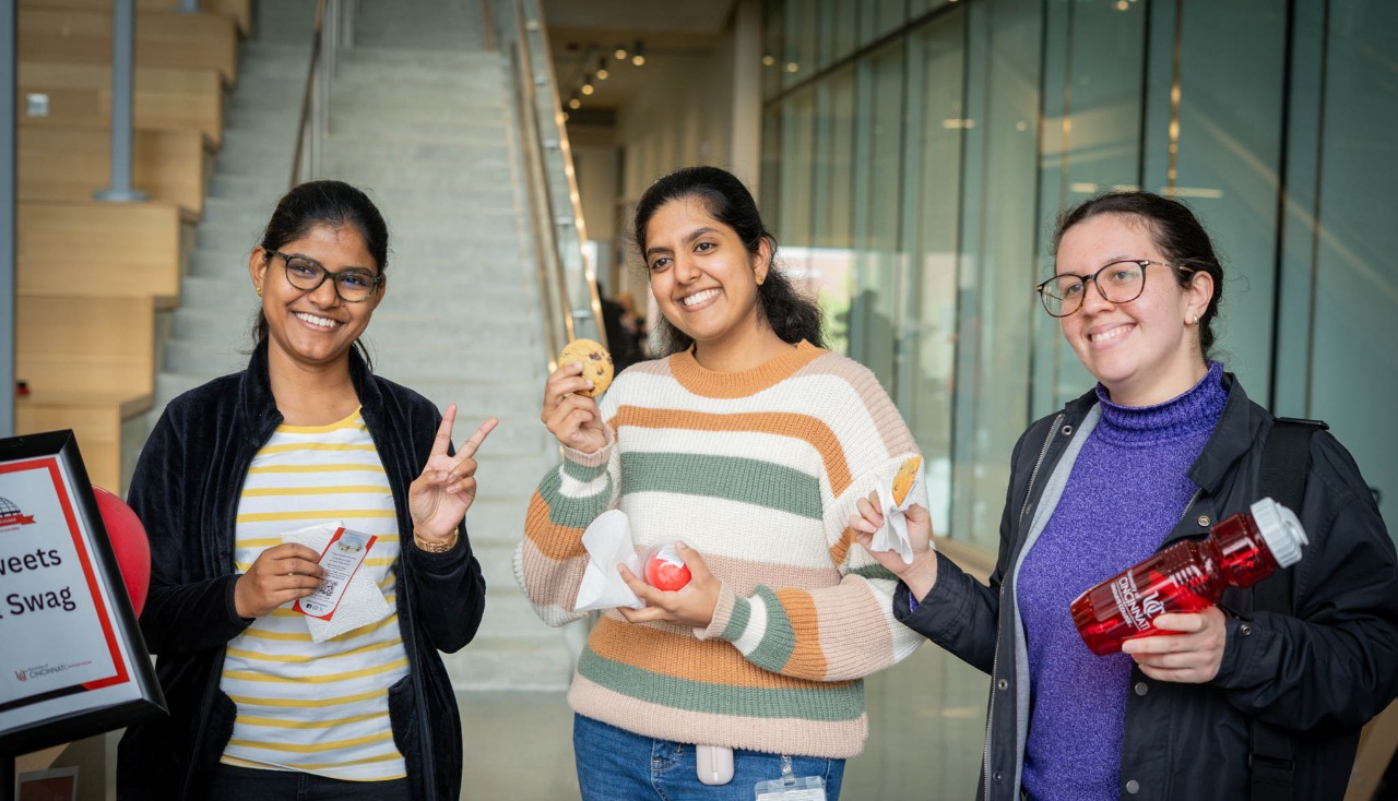 Three smiling students pose on UC Medical Campus holding cookies, Graduate College swag, and snacks.