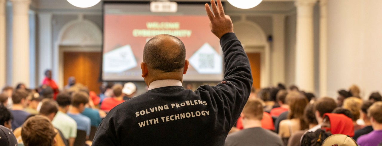 man seen from behind, hand raised, wearing a shirt that reads solving problems with technology