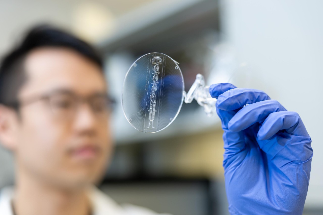 Doctoral Student Heeyong Jang holds up a microfluidic device in a gloved hand.