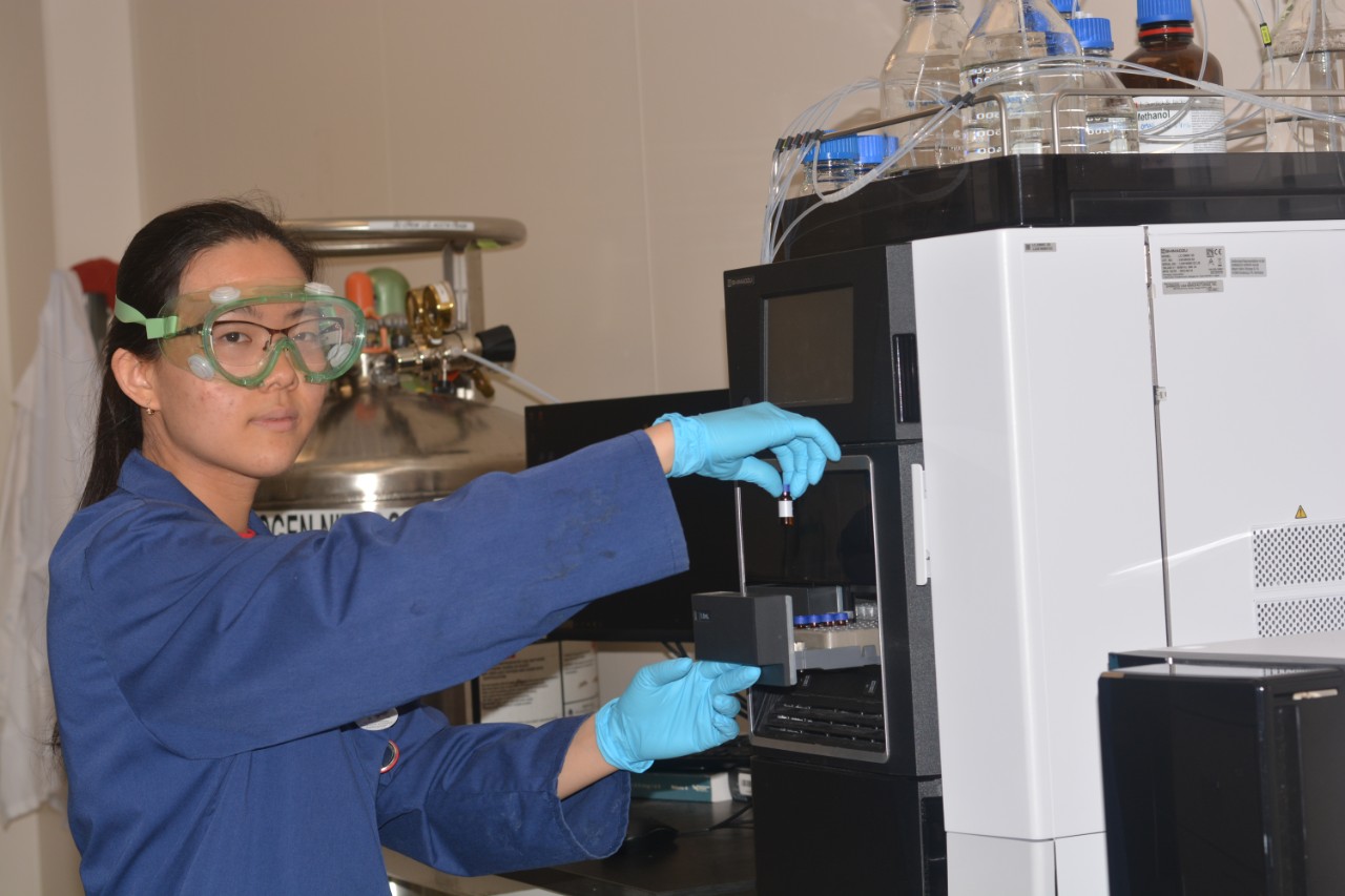 Karen Noda Morishita working in the lab. She holds a small sample by a machine and wears blue rubber gloves, safety goggles, and a blue lab coat. 