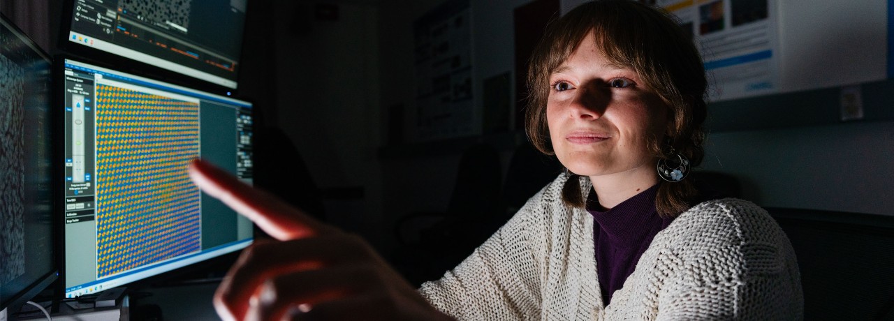 Person pointing at computer screens with data and graphs, in a dimly lit room.