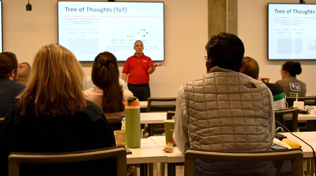 A man in a red shirt and black pants instructs a class full of professionals with laptops open.