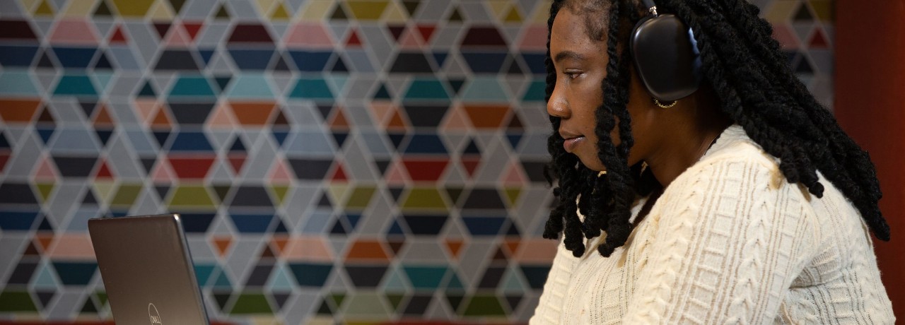 Individual wearing headphones and working at a laptop in an office with a colorful geometric pattern on the wall behind them.