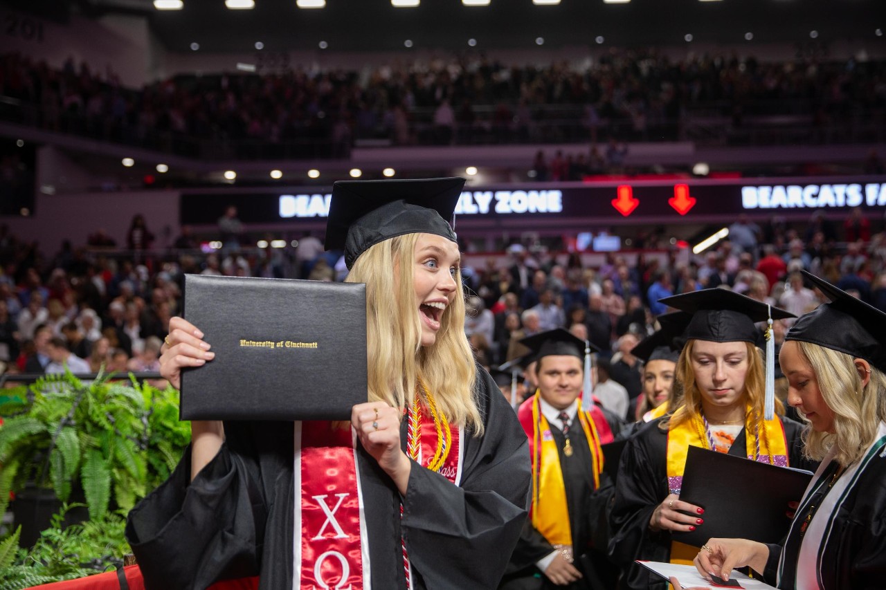 A UC grad in a cap and gown holds up her diploma on the floor of Fifth Third Arena.