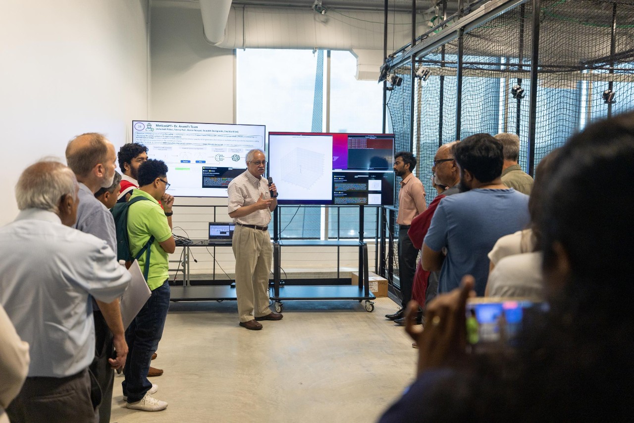 Sam Anand holds a microphone while addressing a group of people who watch TV screens.