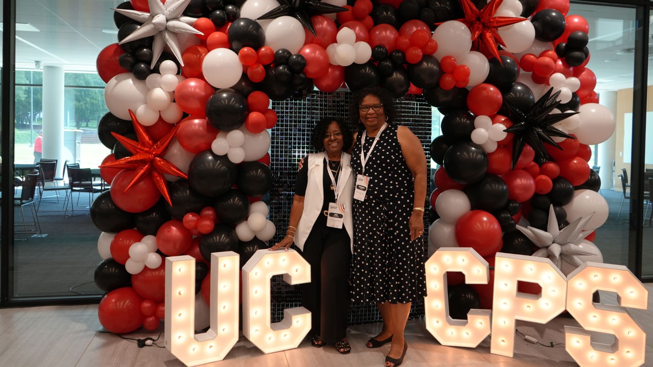 Two people stand with a balloon background and the letters U C and C P S in lights