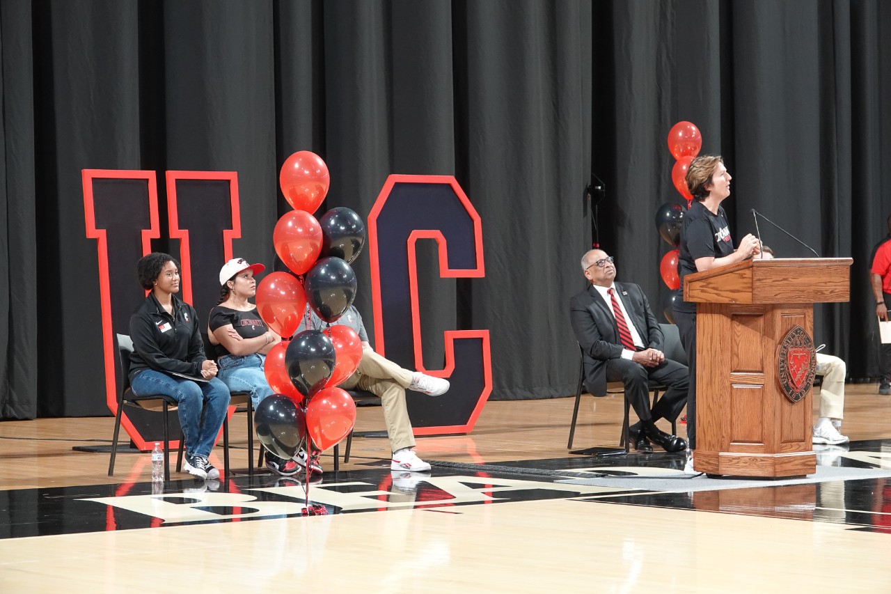 UC officials talk at a podium on the floor of Fifth Third Arena next to a giant UC sign.