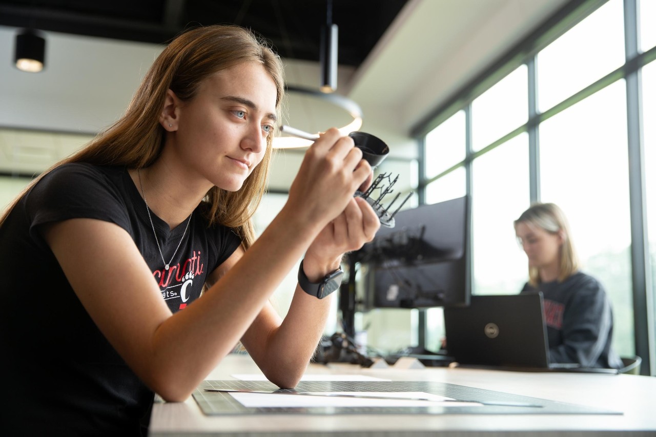 Brooke Boeding shown sitting at work desk with mechanical instrument in hand and female student on a laptop is shown in the background