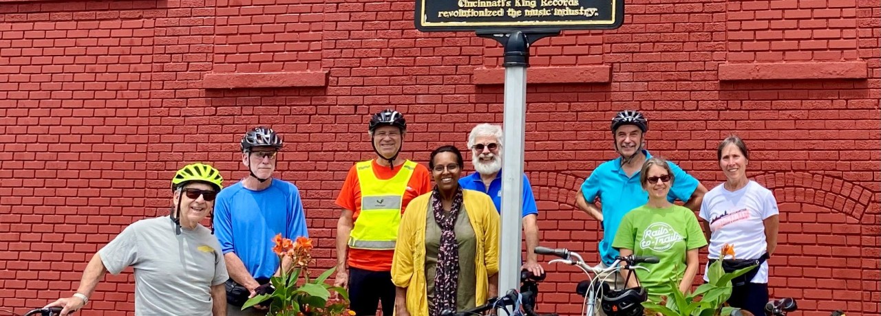 Group of seven cyclists posing with their bikes in front of a red brick wall, under a street sign that reads "King Records".