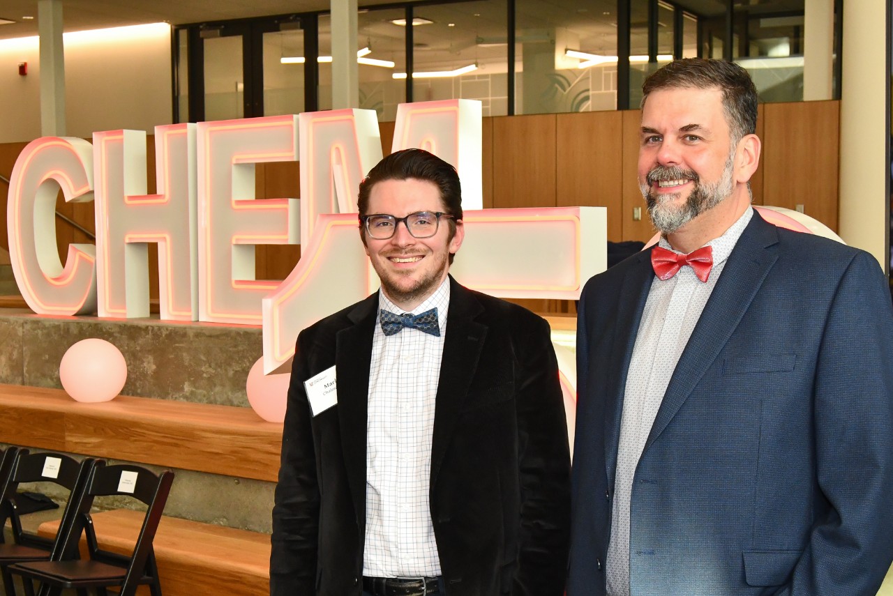 Two people pose in front of a neon Chem 150 sign.