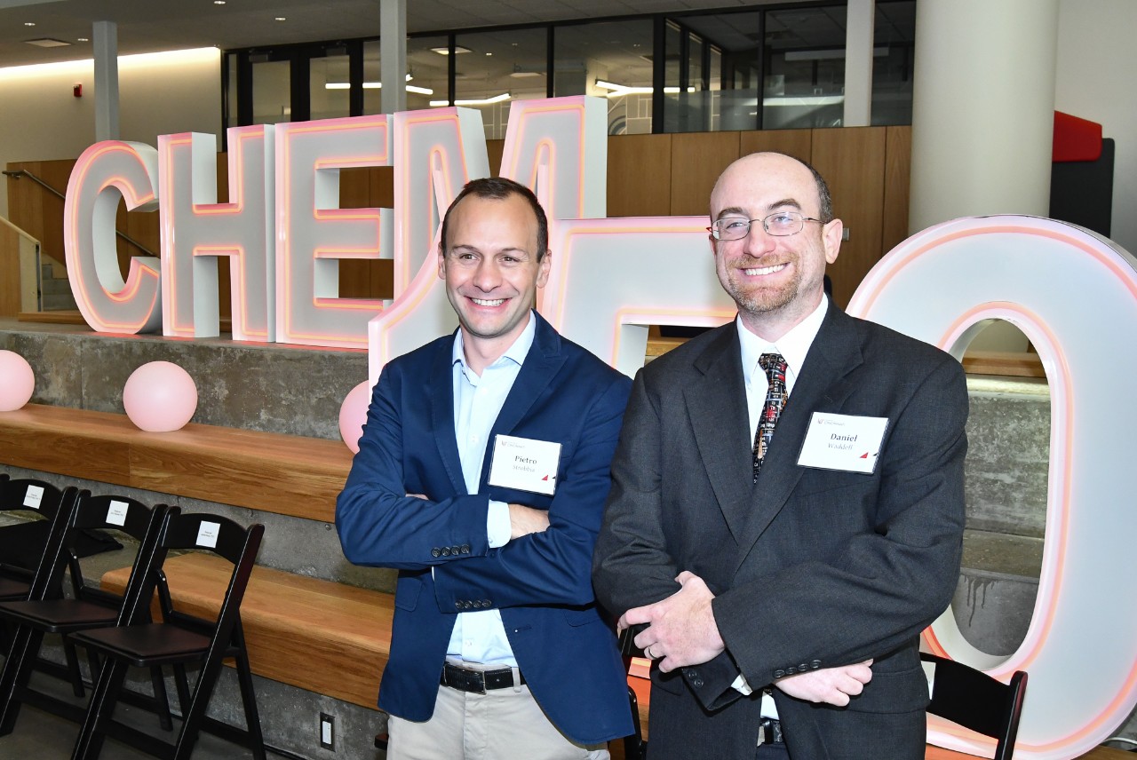Two people stand in front of a neon Chem 150 sign.