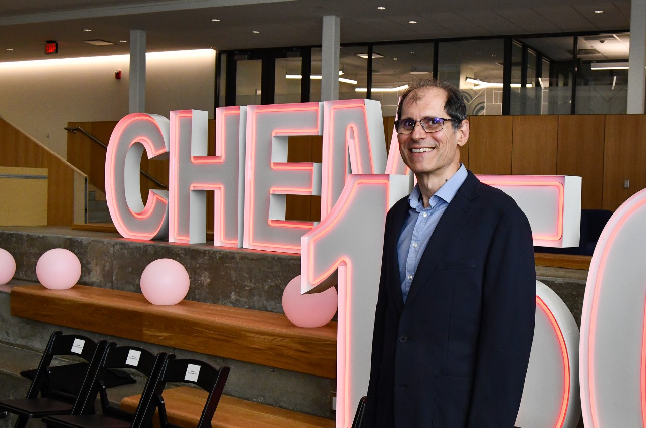 George Stan stands in front of a neon Chem 150 sign.