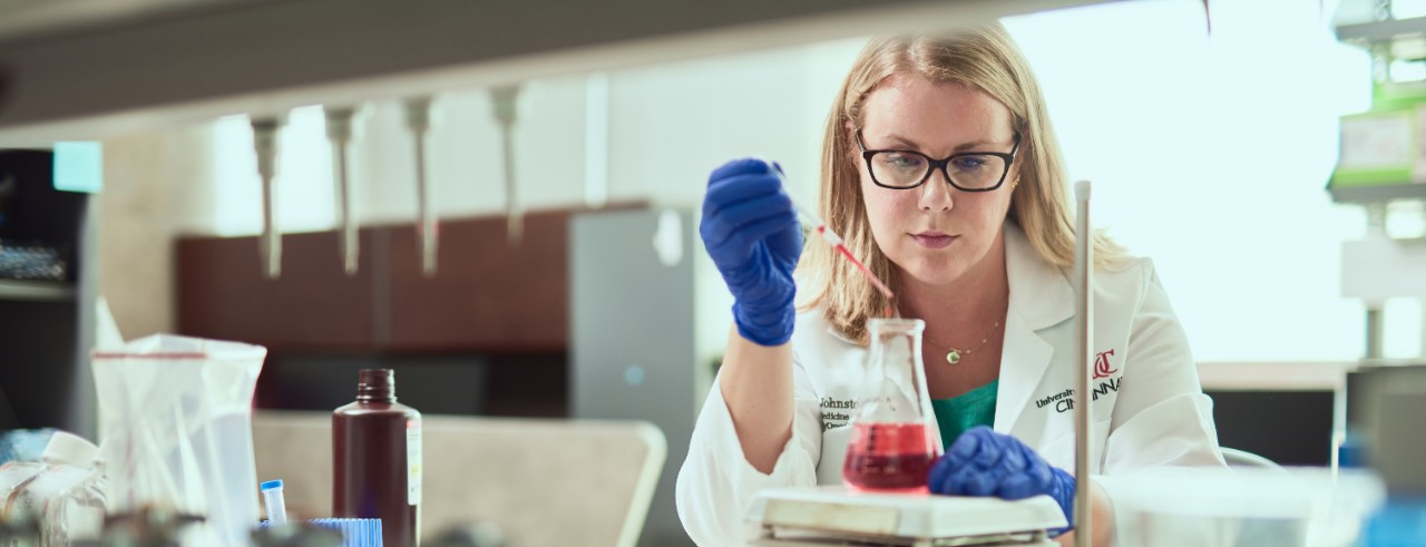 Megan Johnstone, wearing a white lab coat and blue gloves, pipettes a red liquid into a beaker in the lab.