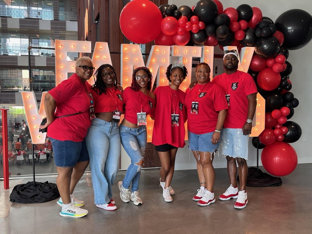 Six people dressed in UC red tops with bearcat logo them pose in front of a Family Weekend light display