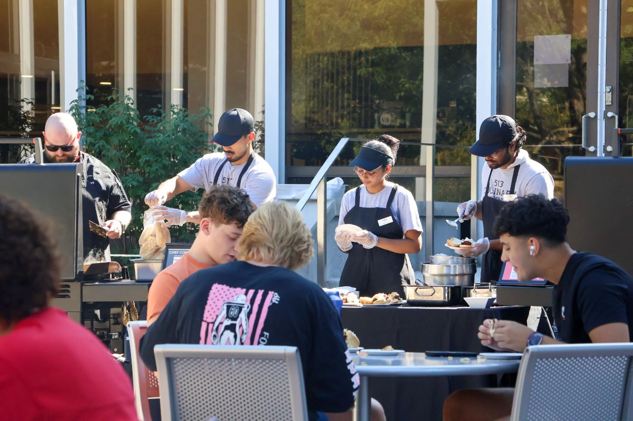 outdoor courtyard with three workers behind a grill and prep area handling food while several people sit at tables out front