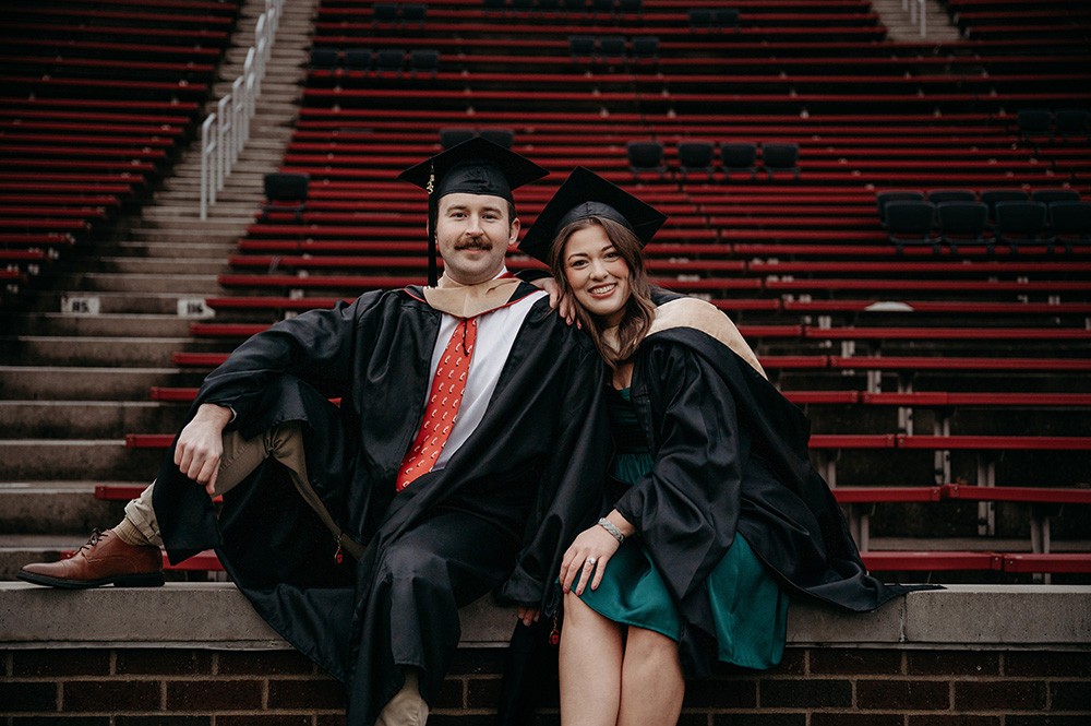 Alanah Jacobson and husband pose in Nippert Stadium in graduation caps and gowns