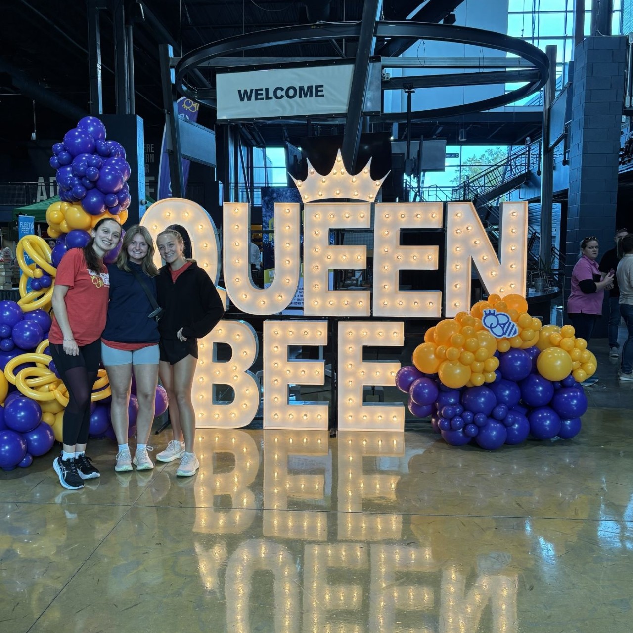 Three female interns standing in front of a sign that says "Queen Bee" and ballons 