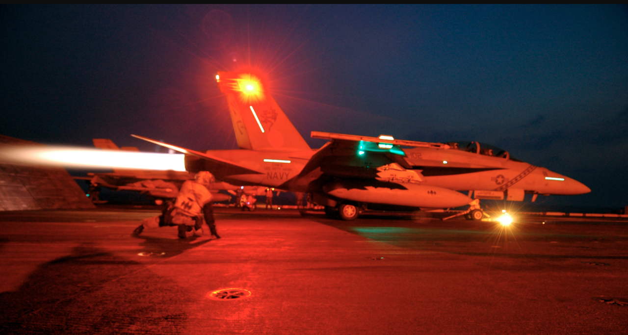 A fighter plane's engines roar as it prepares to take off with a groundcrew member in the foreground on a U.S. aircraft carrier at night.