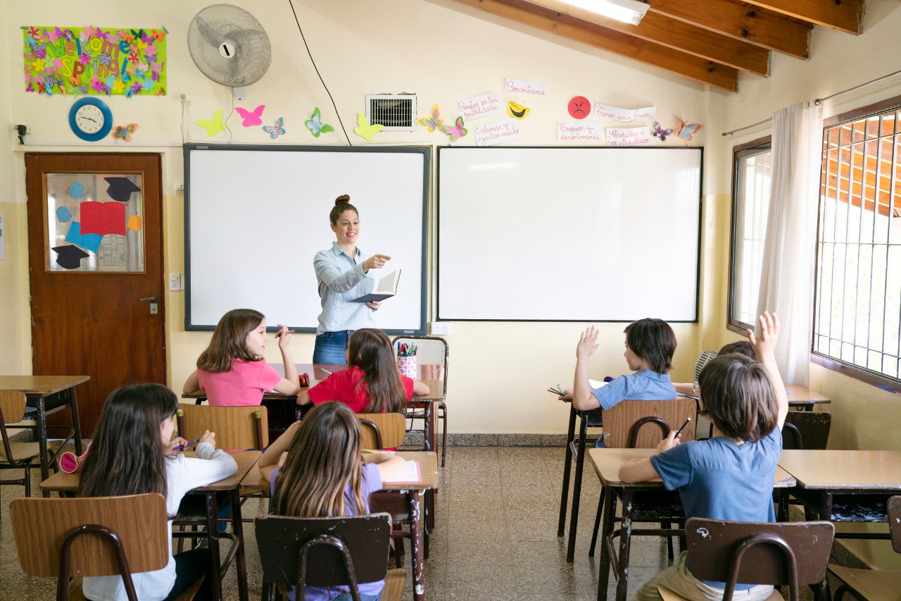 female teacher stands in front of a board in front of a class of four visible students sitting at desks