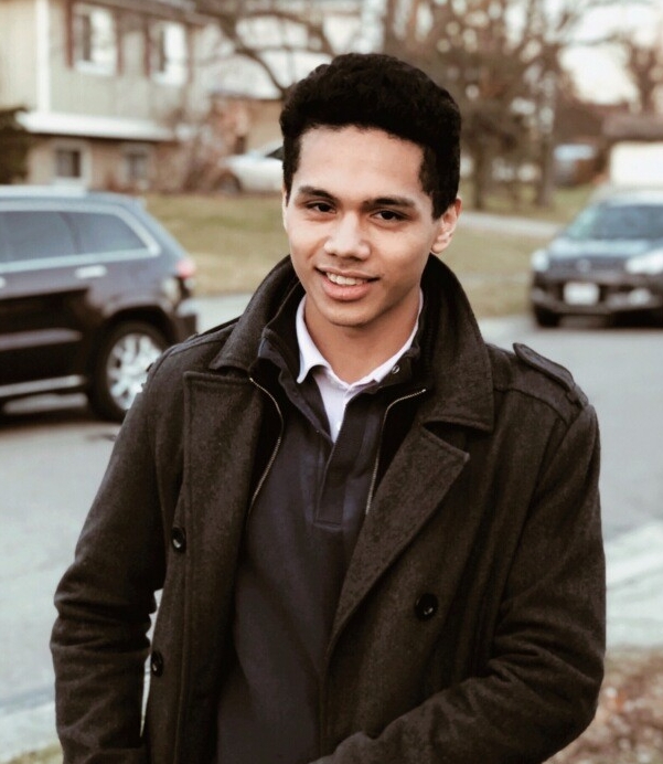 A young man with black hair stands on a sidewalk outside on a bright fall day. He is wearing a brown pea coat and brown sweater and black pants